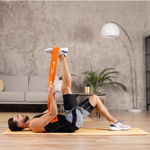 Un homme est allongé sur le dos sur un tapis de sport jaune et tient une bande élastique Hop-Sport orange, qui est accrochée à sa jambe levée haut dans les airs.