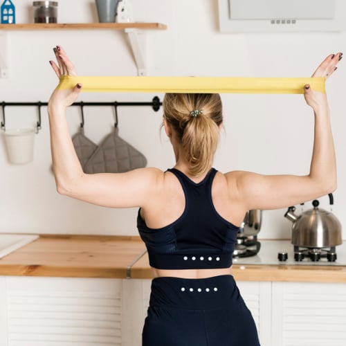 Une femme tournée de dos étire au-dessus de sa tête une bande élastique de musculation jaune