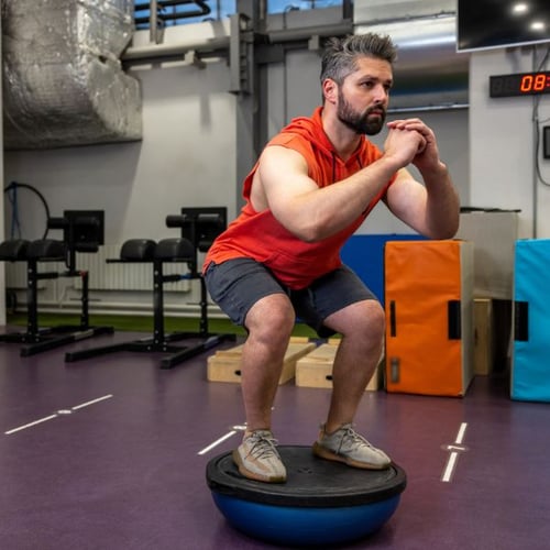 Un homme à la salle de sport se tient debout sur une planche d'équilibre et effectue un squat.