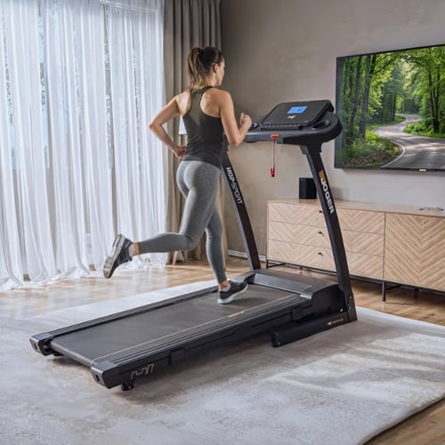 Une femme en leggings gris court sur un tapis de course en regardant la télévision dans le salon