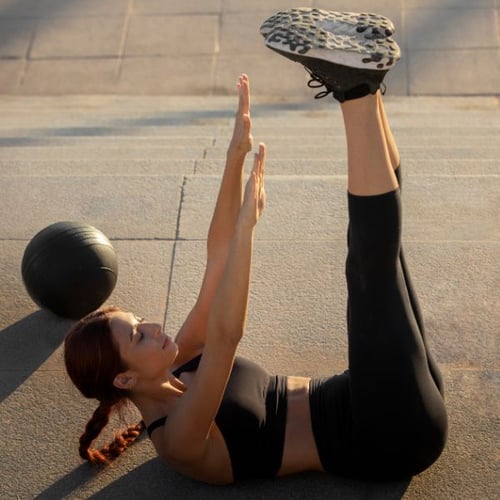 Une femme est allongée à côté d'un ballon d'exercice et lève simultanément les bras et les jambes vers le haut en faisant un hollow hold intense.